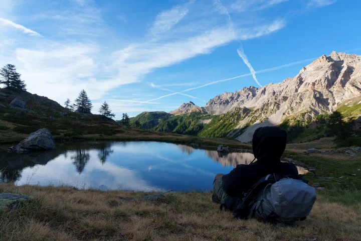 Sitting on the grass beside a still alpine lake in a hooded jacket next to a large backpack, looking out at jagged peaks reflected in the water